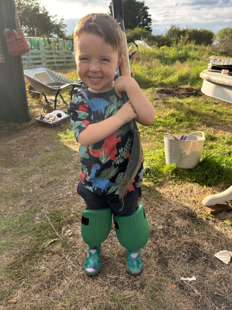 Child on the community allotment Child wearing green welly boots and a colourful shirt, smiling while holding a gardening tool.